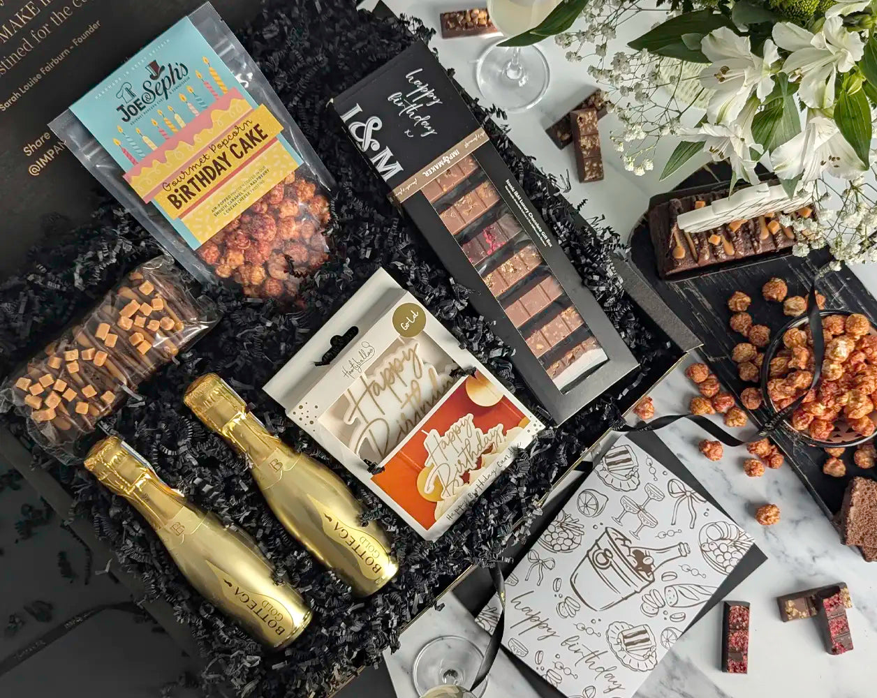 Gourmet gift box with chocolate, cake, candle, popcorn, and gold bottles of Prosecco on a marble surface.