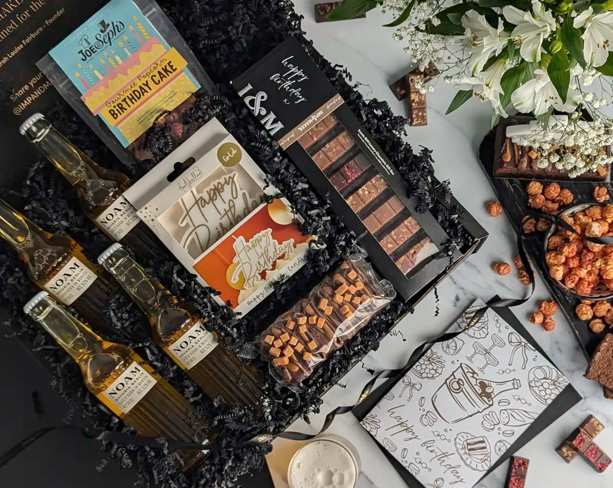 Gourmet gift basket with chocolate bars, mini bottles, and a birthday card on a decorative surface.