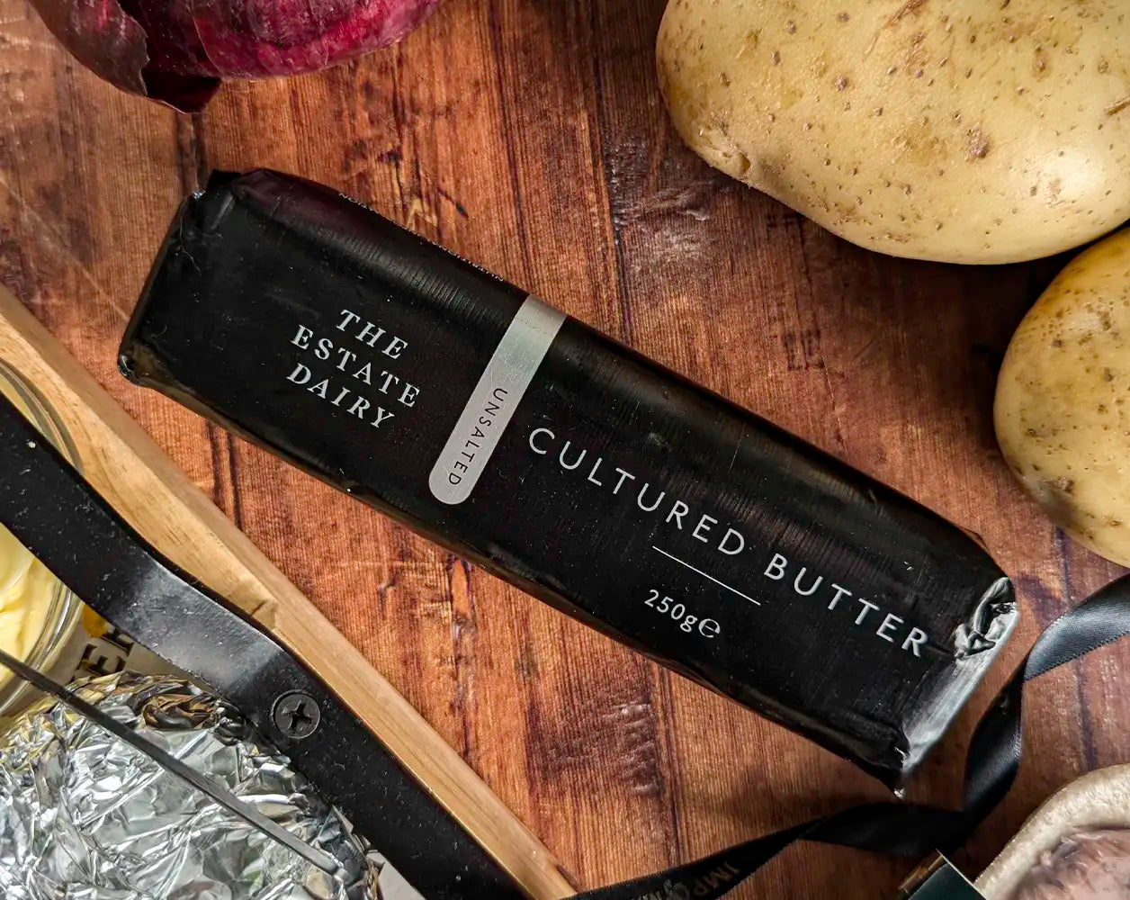 Bar of cultured butter from The Estate Dairy on a wooden surface with potatoes and an onion.