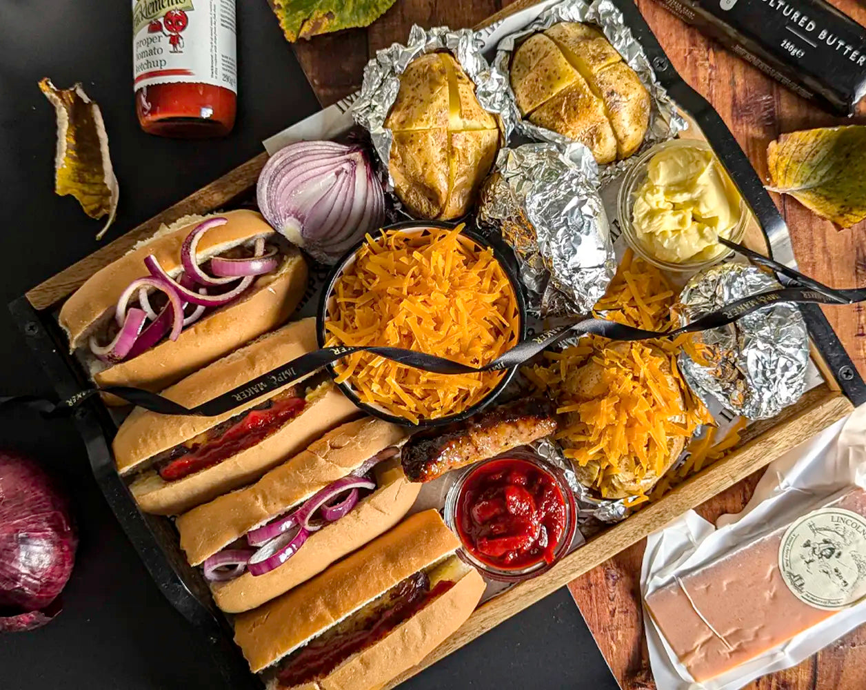 Hot dog platter with buns, baked potatoes, condiments, and sides on a wooden tray.