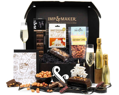 Gourmet gift box with Imp & Maker branding, including snacks, bottles of Prosecco, and a cake, on a white background.