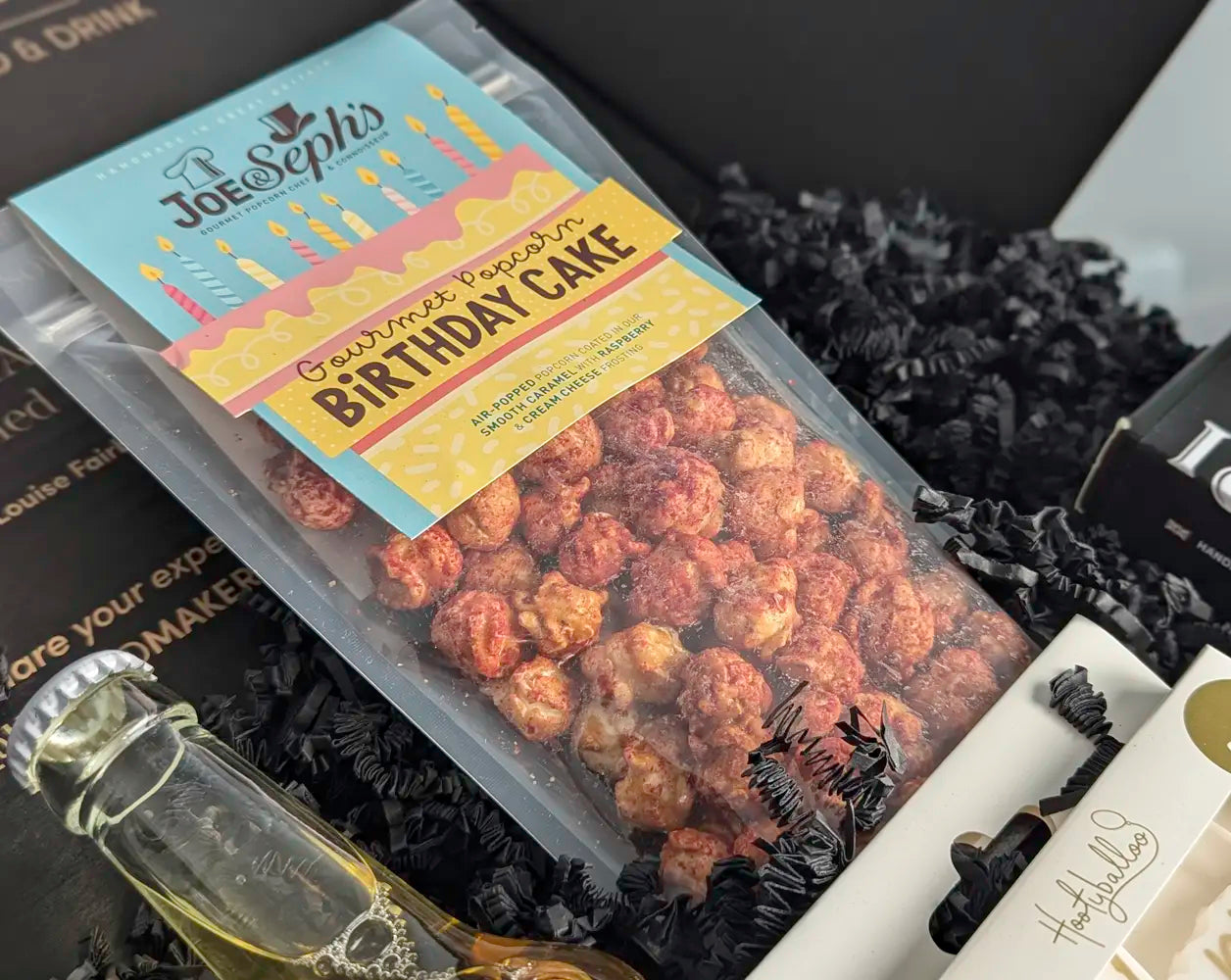 Packaged gourmet birthday cake popcorn with a decorative label on a black textured background.