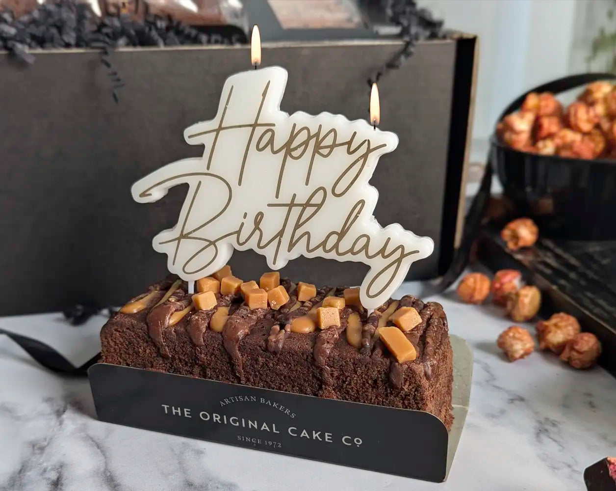 Small birthday cake with 'Happy Birthday' candle and visible brand name on a marble surface.