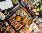 Assorted food items including sausages, cheese, and condiments on a wooden board with a bouquet of flowers.